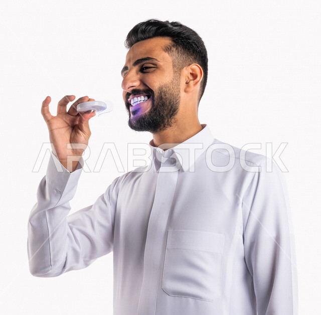 بورتريه بصورة مقربة لرجل عربي خليجي سعودي ، يقوم بإستخدام جهاز تبييض الأسنان المنزلي ، مفهوم العناية والإهتمام بصحة و نظافة الأسنان ، علاجات طب الأسنان ، جلسات العناية بتبييض الأسنان، خلفية بيضاء ، جهاز ليزر