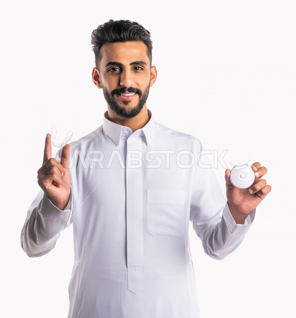 بورتريه بصورة مقربة لرجل عربي خليجي سعودي، يحمل بيده جهاز تبييض الأسنان المنزلي وطقم أسنان بيضاء ، مفهوم العناية والإهتمام بصحة ومظهر الأسنان، علاجات طب الأسنان، جلسات العناية بتبييض الأسنان، خلفية بيضاء، جهاز ليزر