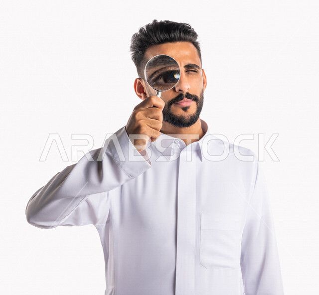 بورتريه بصورة مقربة لرجل عربي خليجي سعودي ، ينظر من خلال العدسة المكبرة ، أدوات بصرية طبية ، الرؤية عن قرب بإستخدام العدسة المكبرة ، خلفية بيضاء
