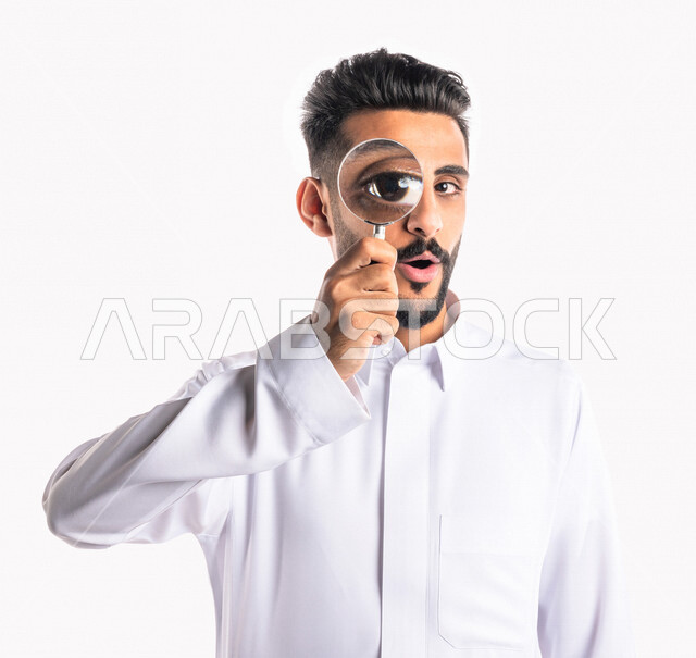 بورتريه بصورة مقربة لرجل عربي خليجي سعودي ، ينظر من خلال العدسة المكبرة ، أدوات بصرية طبية ، الرؤية عن قرب بإستخدام العدسة المكبرة ، خلفية بيضاء
