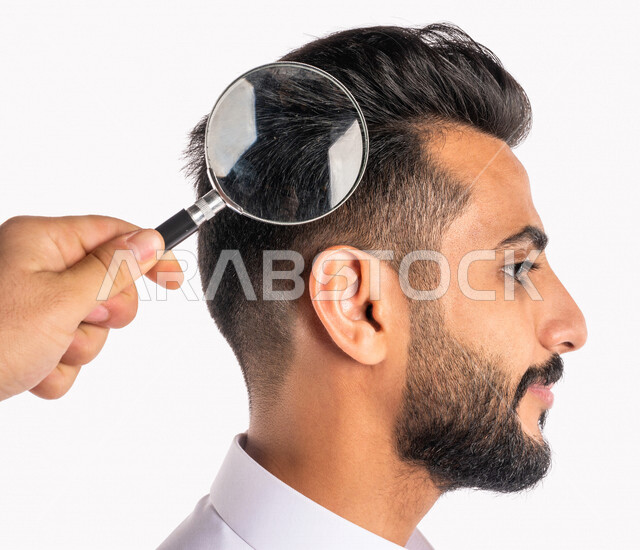 بورتريه بصورة مقربة ليد تحمل عدسة مكبرة ، الرؤية عن قرب بإستخدام العدسة المكبرة على رأس رجل عربي خليجي سعودي ، أدوات بصرية طبية ، فحص فروة رأس من القشرة ، مفهوم النظافة الشخصية ، خلفية بيضاء