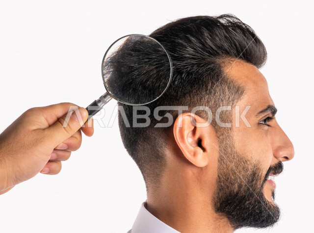 بورتريه بصورة مقربة ليد تحمل عدسة مكبرة ، الرؤية عن قرب بإستخدام العدسة المكبرة على رأس رجل عربي خليجي سعودي ، أدوات بصرية طبية ، فحص فروة رأس من القشرة ، مفهوم النظافة الشخصية ، خلفية بيضاء