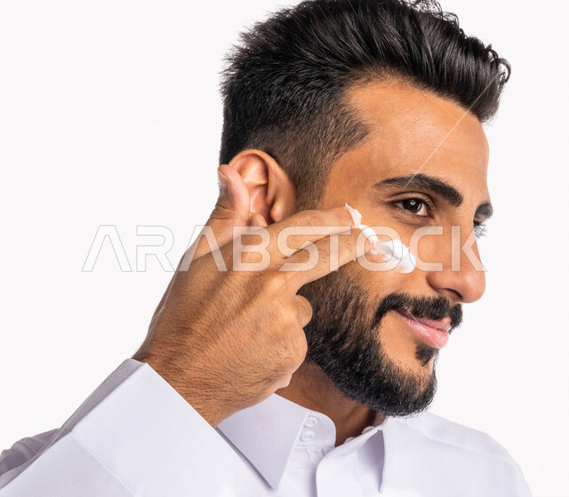 بورتريه بصورة مقربة لرجل عربي خليجي سعودي، يضع كريم الترطيب على بشرة الوجه، مفهوم العناية والإهتمام بنضارة وتحسين البشرة، روتين العناية بالبشرة، خلفية بيضاء، خطوات تنظيف البشرة، ترطيب وتفتيح البشرة، إستخدام مكونات تجميلية، جلسات العناية بالبشرة