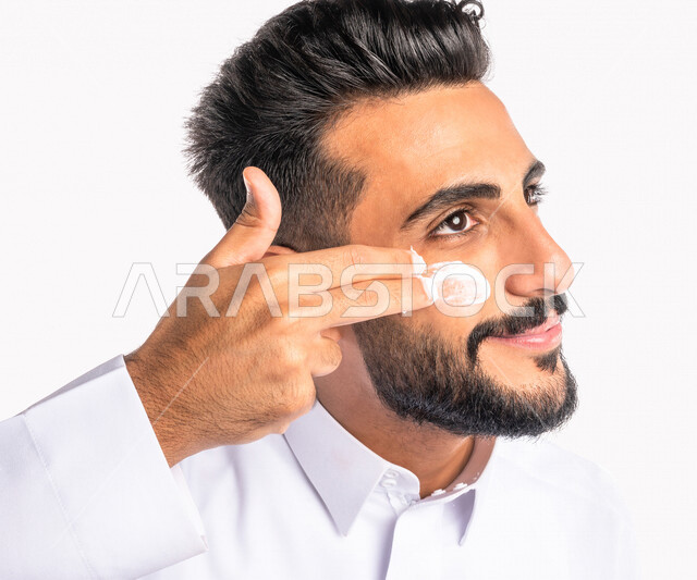 بورتريه بصورة مقربة لرجل عربي خليجي سعودي، يضع كريم الترطيب على بشرة الوجه، مفهوم العناية والإهتمام بنضارة وتحسين البشرة، روتين العناية بالبشرة، خلفية بيضاء، خطوات تنظيف البشرة، ترطيب وتفتيح البشرة، إستخدام مكونات تجميلية، جلسات العناية بالبشرة