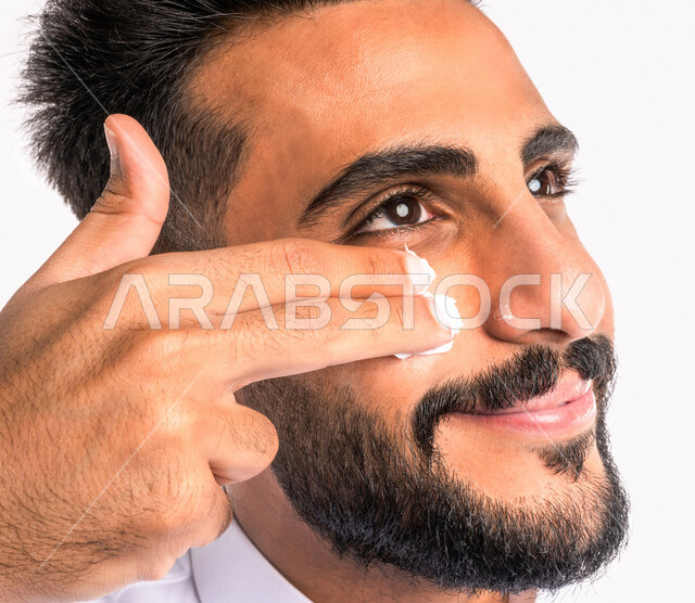 بورتريه بصورة مقربة لرجل عربي خليجي سعودي، يضع كريم الترطيب على بشرة الوجه، مفهوم العناية والإهتمام بنضارة وتحسين البشرة، روتين العناية بالبشرة، خلفية بيضاء، خطوات تنظيف البشرة، ترطيب وتفتيح البشرة، إستخدام مكونات تجميلية، جلسات العناية بالبشرة