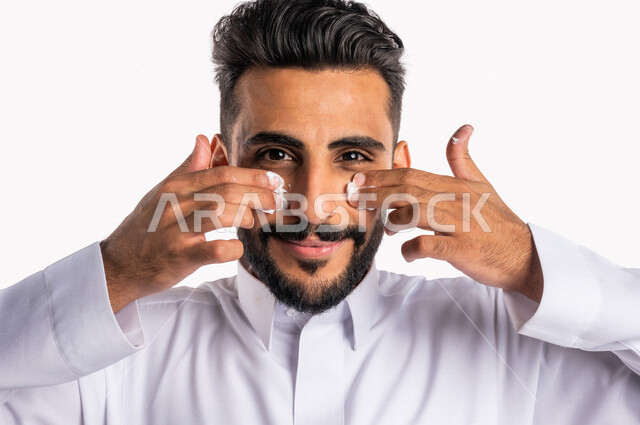 بورتريه بصورة مقربة لرجل عربي خليجي سعودي، يضع كريم الترطيب على بشرة الوجه، مفهوم العناية والإهتمام بنضارة وتحسين البشرة، روتين العناية بالبشرة، خلفية بيضاء، خطوات تنظيف البشرة، ترطيب وتفتيح البشرة، إستخدام مكونات تجميلية، جلسات العناية بالبشرة