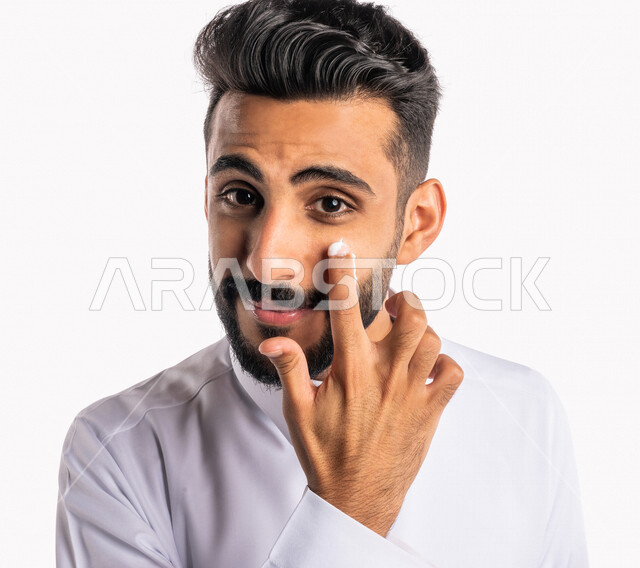 بورتريه بصورة مقربة لرجل عربي خليجي سعودي، يضع كريم الترطيب على بشرة الوجه، مفهوم العناية والإهتمام بنضارة وتحسين البشرة، روتين العناية بالبشرة، خلفية بيضاء، خطوات تنظيف البشرة، ترطيب وتفتيح البشرة، إستخدام مكونات تجميلية، جلسات العناية بالبشرة