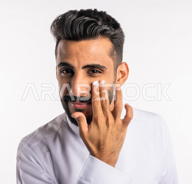 بورتريه بصورة مقربة لرجل عربي خليجي سعودي، يضع كريم الترطيب على بشرة الوجه، مفهوم العناية والإهتمام بنضارة وتحسين البشرة، روتين العناية بالبشرة، خلفية بيضاء، خطوات تنظيف البشرة، ترطيب وتفتيح البشرة، إستخدام مكونات تجميلية، جلسات العناية بالبشرة