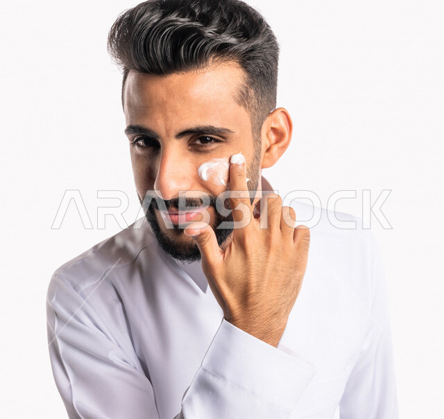 بورتريه بصورة مقربة لرجل عربي خليجي سعودي، يضع كريم الترطيب على بشرة الوجه، مفهوم العناية والإهتمام بنضارة وتحسين البشرة، روتين العناية بالبشرة، خلفية بيضاء، خطوات تنظيف البشرة، ترطيب وتفتيح البشرة، إستخدام مكونات تجميلية، جلسات العناية بالبشرة