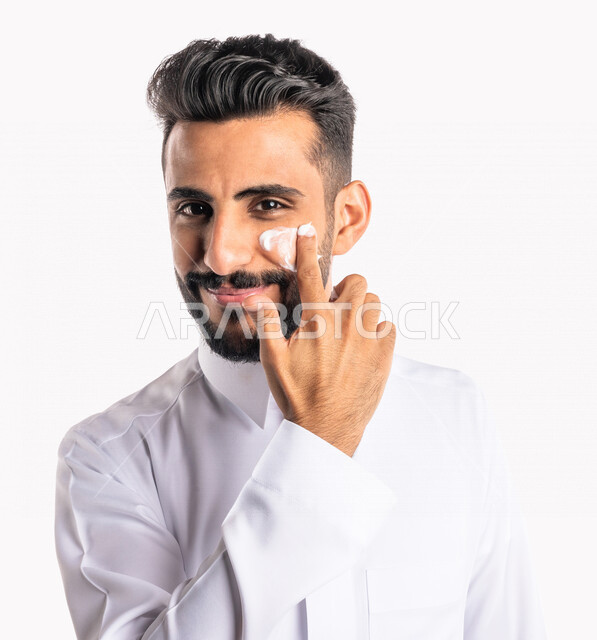 بورتريه بصورة مقربة لرجل عربي خليجي سعودي، يضع كريم الترطيب على بشرة الوجه، مفهوم العناية والإهتمام بنضارة وتحسين البشرة، روتين العناية بالبشرة، خلفية بيضاء، خطوات تنظيف البشرة، ترطيب وتفتيح البشرة، إستخدام مكونات تجميلية، جلسات العناية بالبشرة