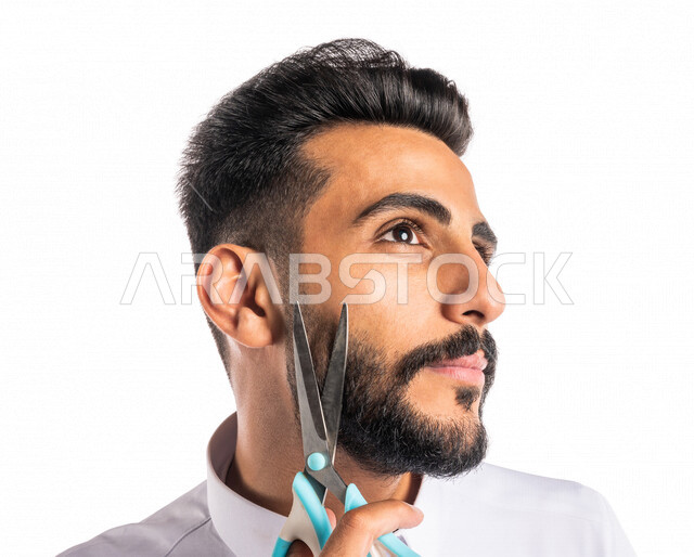 بورتريه بصورة مقربة لرجل عربي خليجي سعودي ، يحمل بيده المقص ، قص و تصفيف الذقن في صالون الحلاقة ، مفهوم العناية و الإهتمام بالجمال والمظهر الخارجي ، صالون حلاقة تجميلي رجالي ، خلفية بيضاء ، العناية والإهتمام بشعر صحي ونظيف