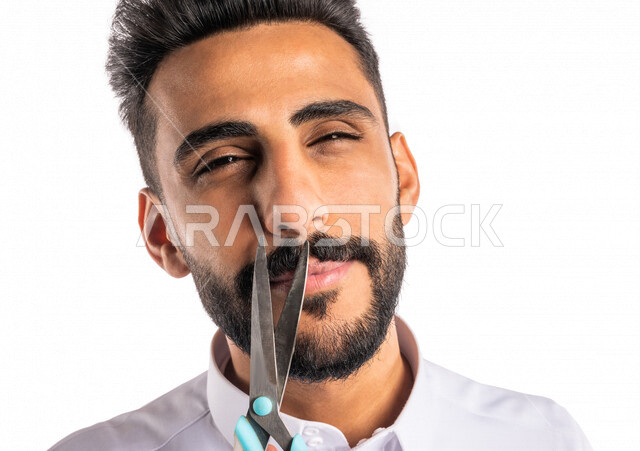 بورتريه بصورة مقربة لرجل عربي خليجي سعودي ، يحمل بيده المقص ، قص و تصفيف الذقن في صالون الحلاقة ، مفهوم العناية و الإهتمام بالجمال والمظهر الخارجي ، صالون حلاقة تجميلي رجالي ، خلفية بيضاء ، العناية والإهتمام بشعر صحي ونظيف