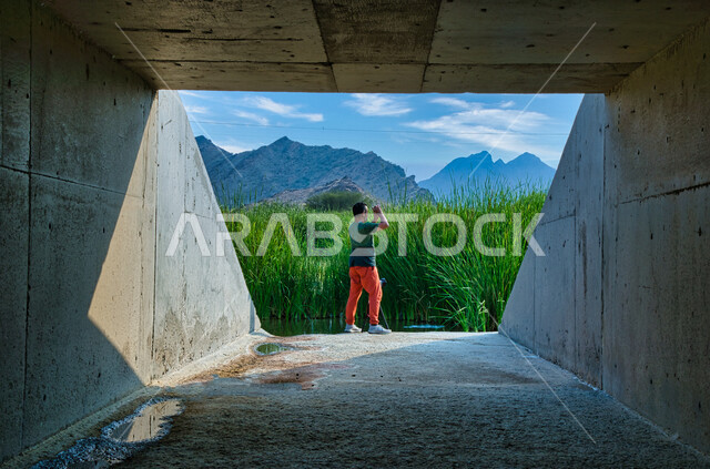 سائح عربي  يقف داخل القناة الخرسانية , منظر جمالي أخاذ , تصوير الطبيعة الخلابة من خلال الهاتف المحمول , الاستمتاع بالطبيعة , رحلة استكشاف الى الطبيعة ,الجبال والسماء الزرقاء الصافية , السياحة في السعودية .  