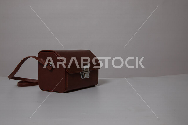 صوره مقربه لحقيبه مصنوعه من الجلد, حقيبه جلديه, حافظة للكاميرا, شنطه نسائيه على طاوله بيضاء, خلفيه بيضاء, حقيبه باللون البني, أزياء و إكسسوارات