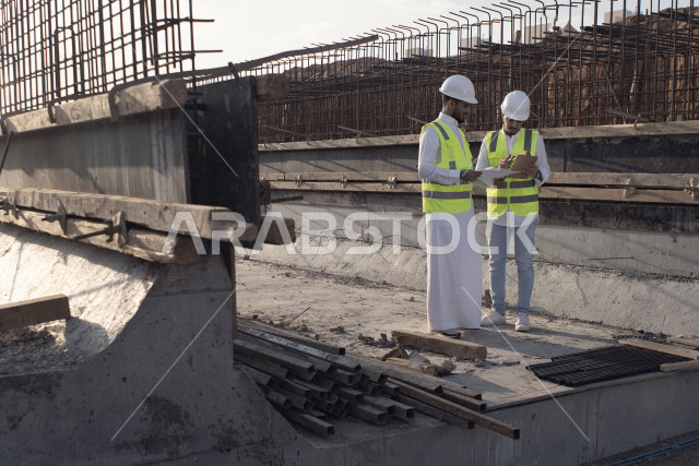 Saudi Arabian Gulf Consulting Engineer meeting with the professional contractor, wearing protective suit and safety helmet, discussing construction blueprint paper, building strategic plans, working on construction site