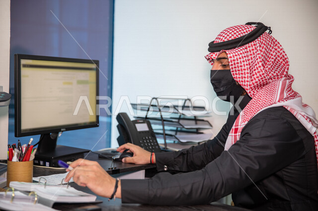 صورة مقربة لشاب عربي خليجي سعودي يرتدي الكمامة الواقية، العمل من خلال الحاسوب، اعمال ومكاتب، بيئة عمل مكتبي، ادخال بيانات، الالتزام باجراءات الوقاية من فايروس كورونا، مهن ووظائف