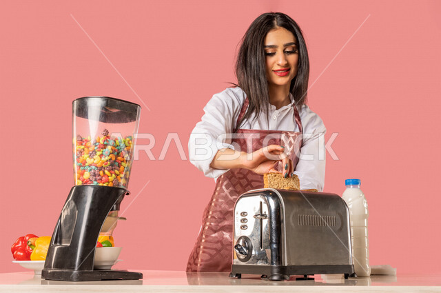 بورتريه لمرأة عربية، تقوم بإعداد الفطور الصباحي على طاولة المطبخ، إتباع نظام غذائي صحي، إعداد شرائح خبز التوست، ربة منزل، مفهوم الأكل الصحي، الإفطار الصحي، وجبة فطور صحية، التدبير المنزلي، خلفية ملونة
