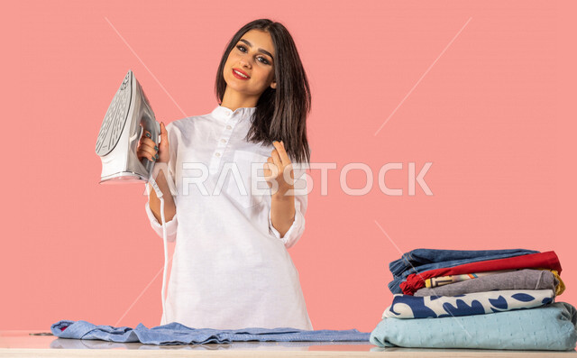 بورتريه لمرأة عربية، تحمل بيدها المكواة الكهربائية، الإستعداد لكي الملابس على لوح الكي، كي وتنظيم الملابس، المهام والأعمال المنزلية الروتينية، أجهزة كهرومنزلية، مفهوم كي وتجفيف الملابس، خلفية ملونة، ربة منزل، التدبير المنزلي