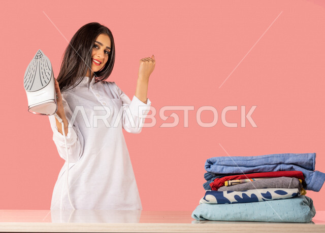 بورتريه لمرأة عربية، تحمل بيدها المكواة الكهربائية، تعابير تدل على الإستعداد لكي الملابس على لوح الكي، كي وتنظيم الملابس، المهام والأعمال المنزلية الروتينية، أجهزة كهرومنزلية، مفهوم كي وتجفيف الملابس، خلفية ملونة، ربة منزل، التدبير المنزلي