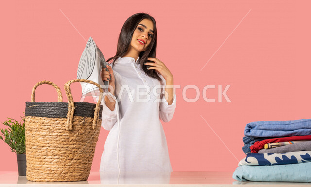 بورتريه لمرأة عربية، تحمل بيدها المكواة الكهربائية، الإستعداد لكي الملابس على لوح الكي، كي وتنظيم الملابس، المهام والأعمال المنزلية الروتينية، أجهزة كهرومنزلية، مفهوم كي وتجفيف الملابس، خلفية ملونة، ربة منزل، التدبير المنزلي