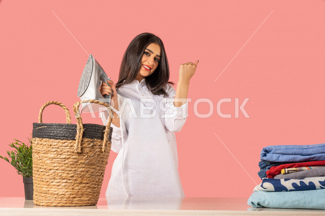 بورتريه لمرأة عربية، تحمل بيدها المكواة الكهربائية، تعابير تدل على الإستعداد لكي الملابس على لوح الكي، كي وتنظيم الملابس، المهام والأعمال المنزلية الروتينية، أجهزة كهرومنزلية، مفهوم كي وتجفيف الملابس، خلفية ملونة، ربة منزل، التدبير المنزلي