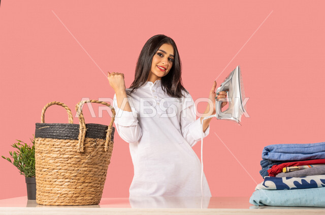 بورتريه لمرأة عربية، تحمل بيدها المكواة الكهربائية، تعابير تدل على الإستعداد لكي الملابس على لوح الكي، كي وتنظيم الملابس، المهام والأعمال المنزلية الروتينية، أجهزة كهرومنزلية، مفهوم كي وتجفيف الملابس، خلفية ملونة، ربة منزل، التدبير المنزلي