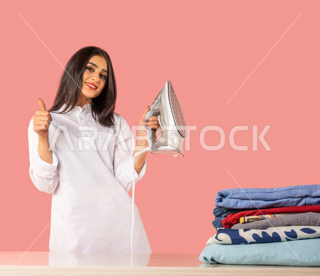 بورتريه لمرأة عربية، تحمل بيدها المكواة الكهربائية، تعابير تدل على الإعجاب والخدمات الجيدة، الإستعداد لكي الملابس على لوح الكي، كي وتنظيم الملابس، المهام والأعمال المنزلية الروتينية، أجهزة كهرومنزلية، كي وتجفيف الملابس، خلفية ملونة، ربة منزل، التدبير المن