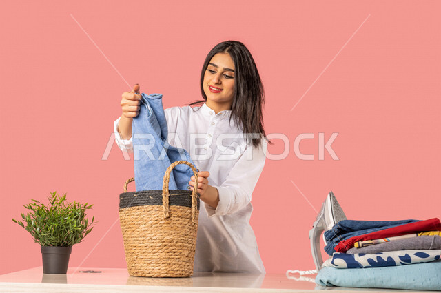 Portrait of a Saudi Arabian Gulf woman, holding clothes in her hand, getting ready to iron clothes using an electric iron, ironing and organizing clothes, routine household tasks and chores, home electrical appliances, the concept of ironing and drying cl