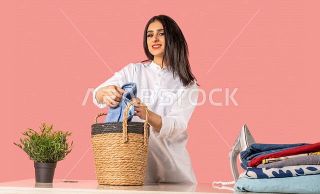 Portrait of a Saudi Arabian Gulf woman, holding clothes in her hand, getting ready to iron clothes using an electric iron, ironing and organizing clothes, routine household tasks and chores, home electrical appliances, the concept of ironing and drying cl