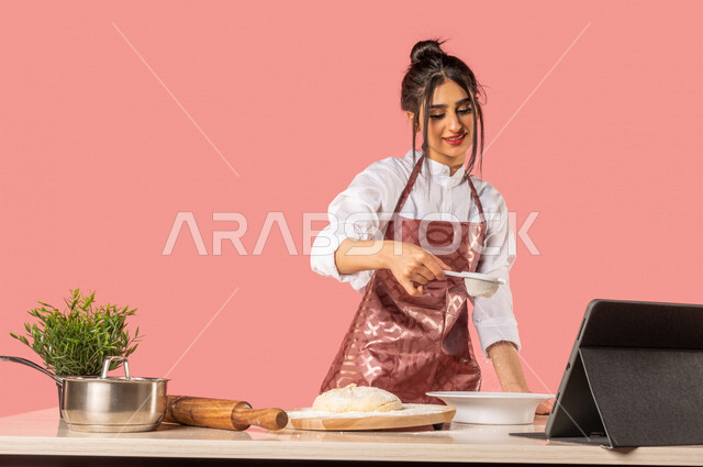 بورتريه لمرأة عربية خليجية سعودية، تقف أمام طاولة عليها مكونات خاصة بالعجين وأدوات خاصة بالمطبخ والعجين، إعداد المخبوزات والمعجنات الشهية، إستخدام جهاز التابلت التقني، طاهية عربية، مشاركة تحضير الوصفات على مواقع التواصل الاجتماعي، ربة منزل