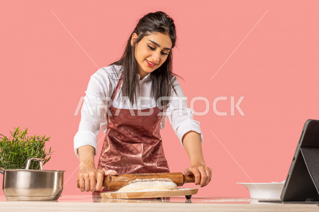 Portrait of a Saudi Arabian Gulf woman preparing Arabic baked goods, using a hand rolling pin (rolling pin), sharing recipes on social media using a technical tablet, a housewife, an Arab chef, a colored background