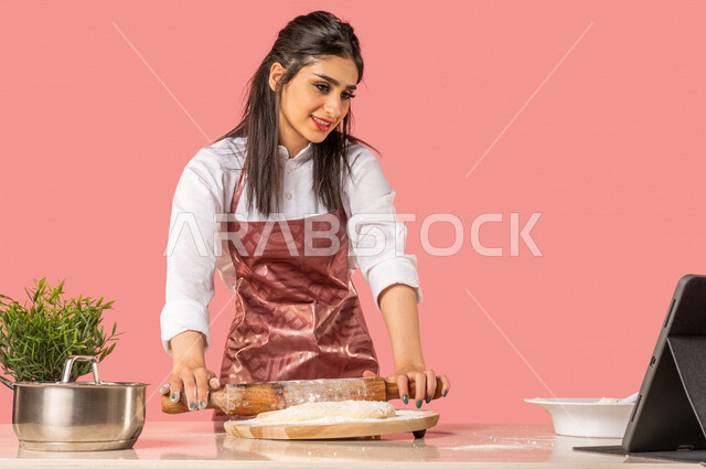 Portrait of a Saudi Arabian Gulf woman preparing Arabic baked goods, using a hand rolling pin (rolling pin), sharing recipes on social media using a technical tablet, a housewife, an Arab chef, a colored background