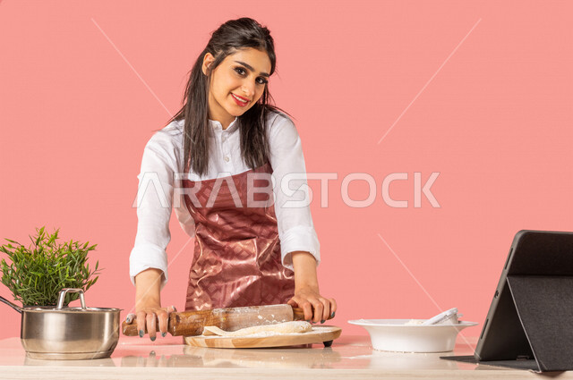 Portrait of a Saudi Arabian Gulf woman preparing Arabic baked goods, using a hand rolling pin (rolling pin), sharing recipes on social media using a technical tablet, a housewife, an Arab chef, a colored background