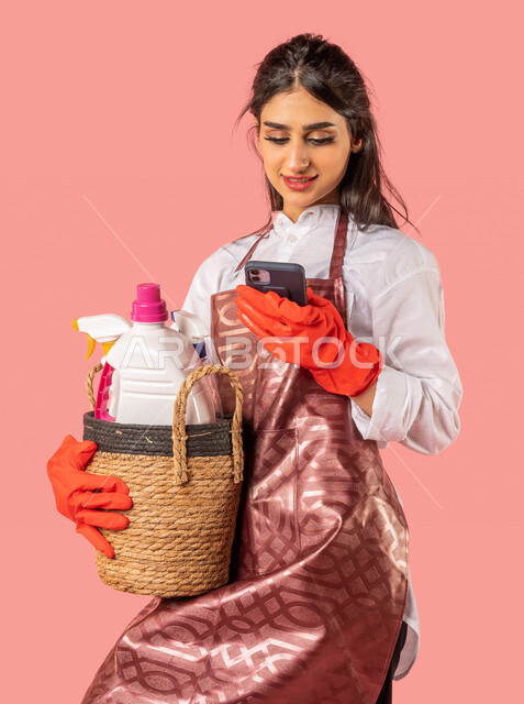 Portrait of an Arab woman wearing an apron and gloves, using a mobile phone, surfing the Internet, holding a straw basket with house cleaning tools, routine housework and tasks, household cleaning tools, cleaning and taking care of housework, housewife, h