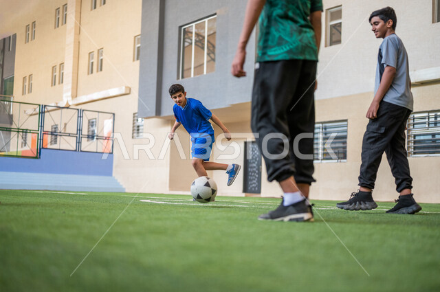 طلاب عرب خليجيين سعوديين يلعبون كرة القدم في ملعب المدرسة، ممارسة التمارين الرياضية، لعب كرة القدم، المراوغة والتحكم بالكرة، حصة التربية الرياضية، التعليم في المملكة العربية السعودية