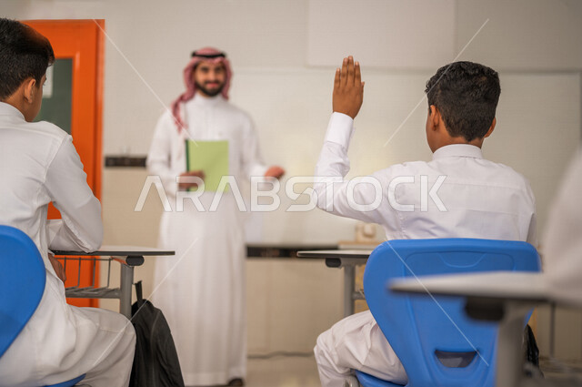 معلم عربي خليجي سعودي يقوم بشرح الدرس، طالب يتفاعل في الحصة الدراسية من خلال رفع يده للاجابة على اسئلة المدرس، المشاركة الفعالة للطلاب، أهداف التربية والتعليم، التفاعل والمشاركة داخل الفصل الدراسي، التعليم في السعودية