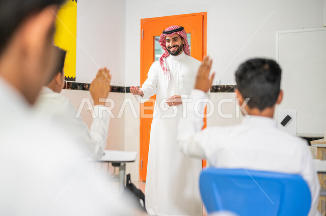 معلم عربي خليجي سعودي يقوم بشرح الدرس، مجموعة من الطلاب يتفاعلون في الحصة الدراسية من خلال رفع ايديهم للاجابة على اسئلة المدرس، المشاركة الفعالة للطلاب، أهداف التربية والتعليم، التفاعل والمشاركة داخل الفصل الدراسي، التعليم في السعودية