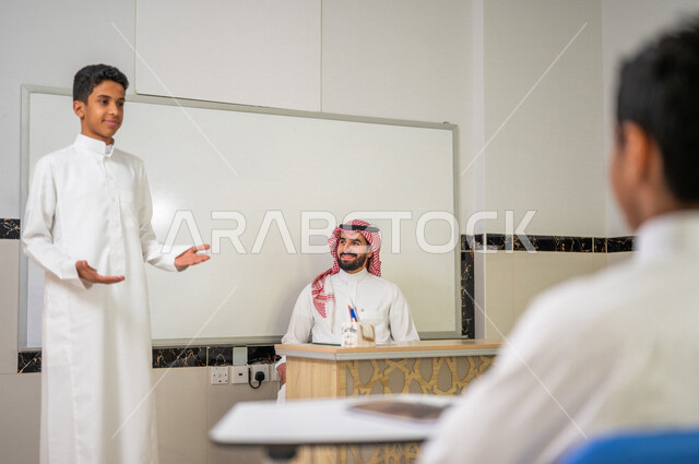 معلم عربي خليجي سعودي يجلس وخلفه لوح مدرسي ابيض، طالب يقوم بشرح الدرس، المشاركة الفعالة للطلاب، التعليم الفعال، أهداف التربية والتعليم، التفاعل والمشاركة داخل الفصل الدراسي، التعليم في السعودية