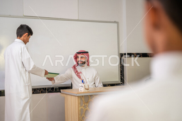 معلم عربي خليجي سعودي يجلس على الكرسي بايماءات وجه مبتسمة، لوحة مدرسية بيضاء، طالب يقوم باعطاء الدفتر للمدرس، المشاركة الفعالة للطلاب، التعليم الفعال، أهداف التربية والتعليم، التفاعل والمشاركة داخل الفصل الدراسي، التعليم في السعودية