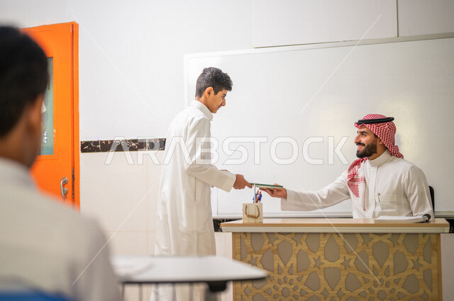 معلم عربي خليجي سعودي يجلس على الكرسي بايماءات وجه مبتسمة، لوحة مدرسية بيضاء، طالب يقوم باعطاء الدفتر للمدرس، المشاركة الفعالة للطلاب، التعليم الفعال، أهداف التربية والتعليم، التفاعل والمشاركة داخل الفصل الدراسي، التعليم في السعودية