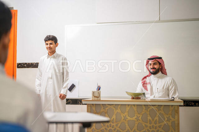 معلم عربي خليجي سعودي يجلس وخلفه لوح مدرسي ابيض، طالب يقوم بشرح الدرس، المشاركة الفعالة للطلاب، التعليم الفعال، أهداف التربية والتعليم، التفاعل والمشاركة داخل الفصل الدراسي، التعليم في السعودية