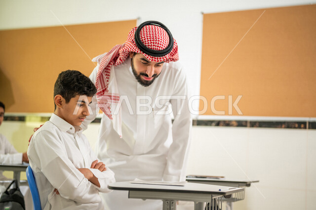 صورة مقربة لطالب عربي خليجي سعودي بايماءات وجه تدل على الحيرة، معلم يقوم بمساعدة الطالب على حل اسئلة الدرس، المشاركة الفعالة للطلاب، التعليم الفعال، أهداف التربية والتعليم، التعليم في السعودية