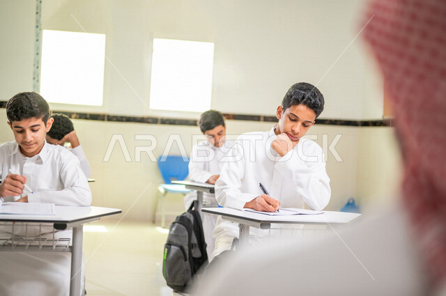 صورة مقربة لطالب عربي خليجي سعودي بايماءات وجه ويدين تدل على التفكير والحيرة، مجموعة من الطلاب يقومون بحل اسئلة الدرس، التمعن والتركيز، المشاركة الفعالة للطلاب، التعليم الفعال، أهداف التربية والتعليم، التعليم في السعودية