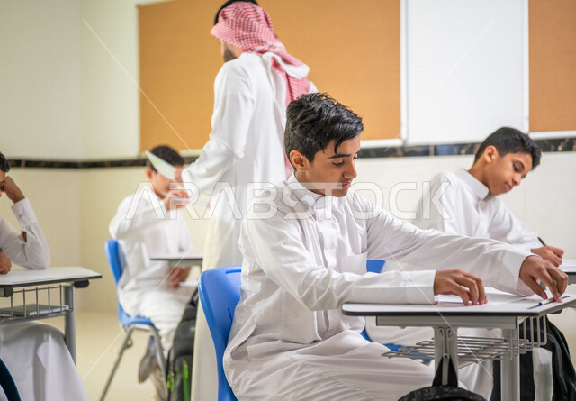 معلم عربي خليجي سعودي يقوم بتوزيع أوراق الاختبارات، طلاب يقومون بحل أسئلة الاختبارات، مراقبة الطلاب اثناء تقديم الاختبارات،  أهداف التربية والتعليم، اختبارات نهائية، نهاية الفصل الدراسي، التعليم في المملكة العربية السعودية