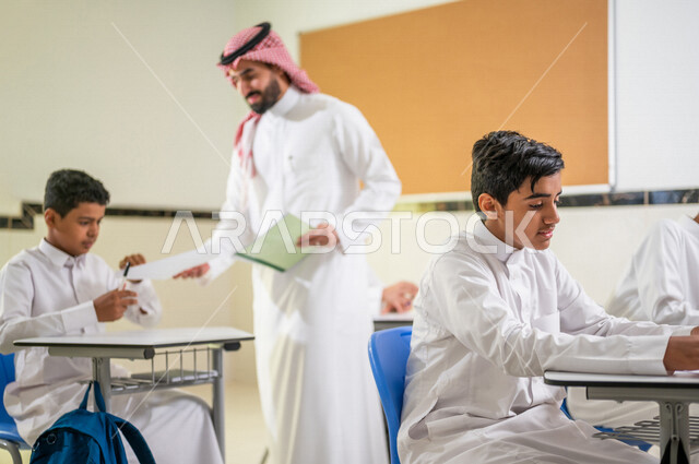معلم عربي خليجي سعودي يقوم بتوزيع أوراق الاختبارات، طلاب يقومون بحل أسئلة الاختبارات، مراقبة الطلاب اثناء تقديم الاختبارات،  أهداف التربية والتعليم، اختبارات نهائية، نهاية الفصل الدراسي، التعليم في المملكة العربية السعودية