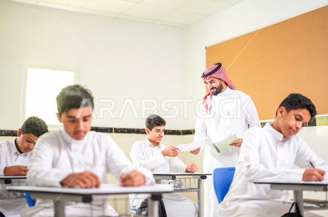 معلم عربي خليجي سعودي يقوم بتوزيع أوراق الاختبارات، طلاب يقومون بحل أسئلة الاختبارات، مراقبة الطلاب اثناء تقديم الاختبارات،  أهداف التربية والتعليم، اختبارات نهائية، نهاية الفصل الدراسي، التعليم في المملكة العربية السعودية