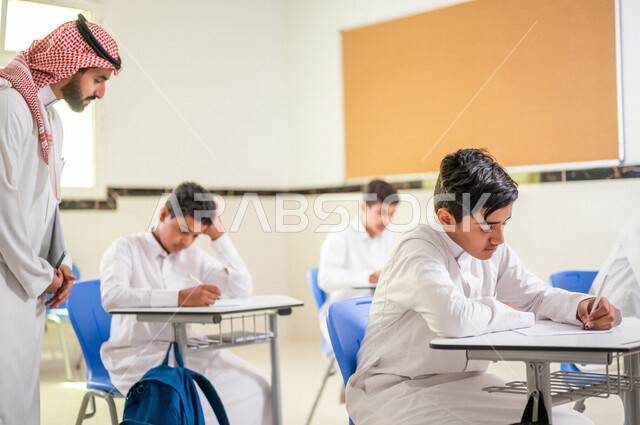 معلم عربي خليجي سعودي يقوم بتوزيع أوراق الاختبارات، طلاب يقومون بحل أسئلة الاختبارات، مراقبة الطلاب اثناء تقديم الاختبارات،  أهداف التربية والتعليم، اختبارات نهائية، نهاية الفصل الدراسي، التعليم في المملكة العربية السعودية