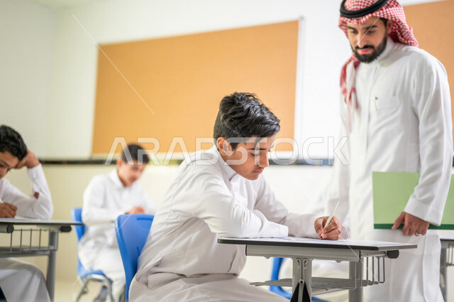 معلم عربي خليجي سعودي يقوم بتوزيع أوراق الاختبارات، طلاب يقومون بحل أسئلة الاختبارات، مراقبة الطلاب اثناء تقديم الاختبارات،  أهداف التربية والتعليم، اختبارات نهائية، نهاية الفصل الدراسي، التعليم في المملكة العربية السعودية