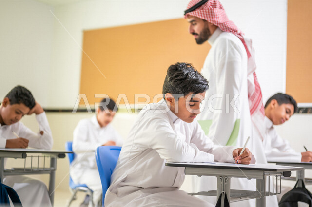 معلم عربي خليجي سعودي يقوم بتوزيع أوراق الاختبارات، طلاب يقومون بحل أسئلة الاختبارات، مراقبة الطلاب اثناء تقديم الاختبارات،  أهداف التربية والتعليم، اختبارات نهائية، نهاية الفصل الدراسي، التعليم في المملكة العربية السعودية