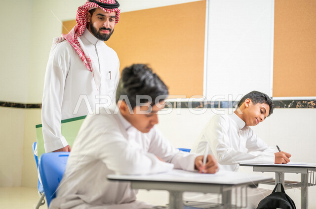 معلم عربي خليجي سعودي يقوم بتوزيع أوراق الاختبارات، طلاب يقومون بحل أسئلة الاختبارات، مراقبة الطلاب اثناء تقديم الاختبارات،  أهداف التربية والتعليم، اختبارات نهائية، نهاية الفصل الدراسي، التعليم في المملكة العربية السعودية